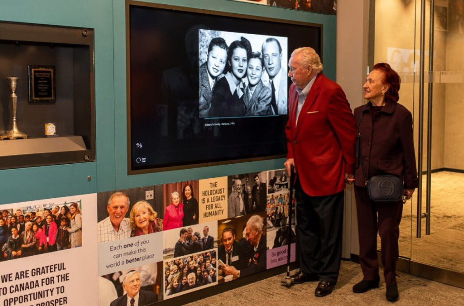 Two older adults stand in front of a museum exhibit screen, projecting a black and white photo.